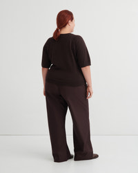 A model with red hair tied back is shown from behind wearing a dark brown short-sleeve top and matching wide-leg trousers against a light gray studio background.