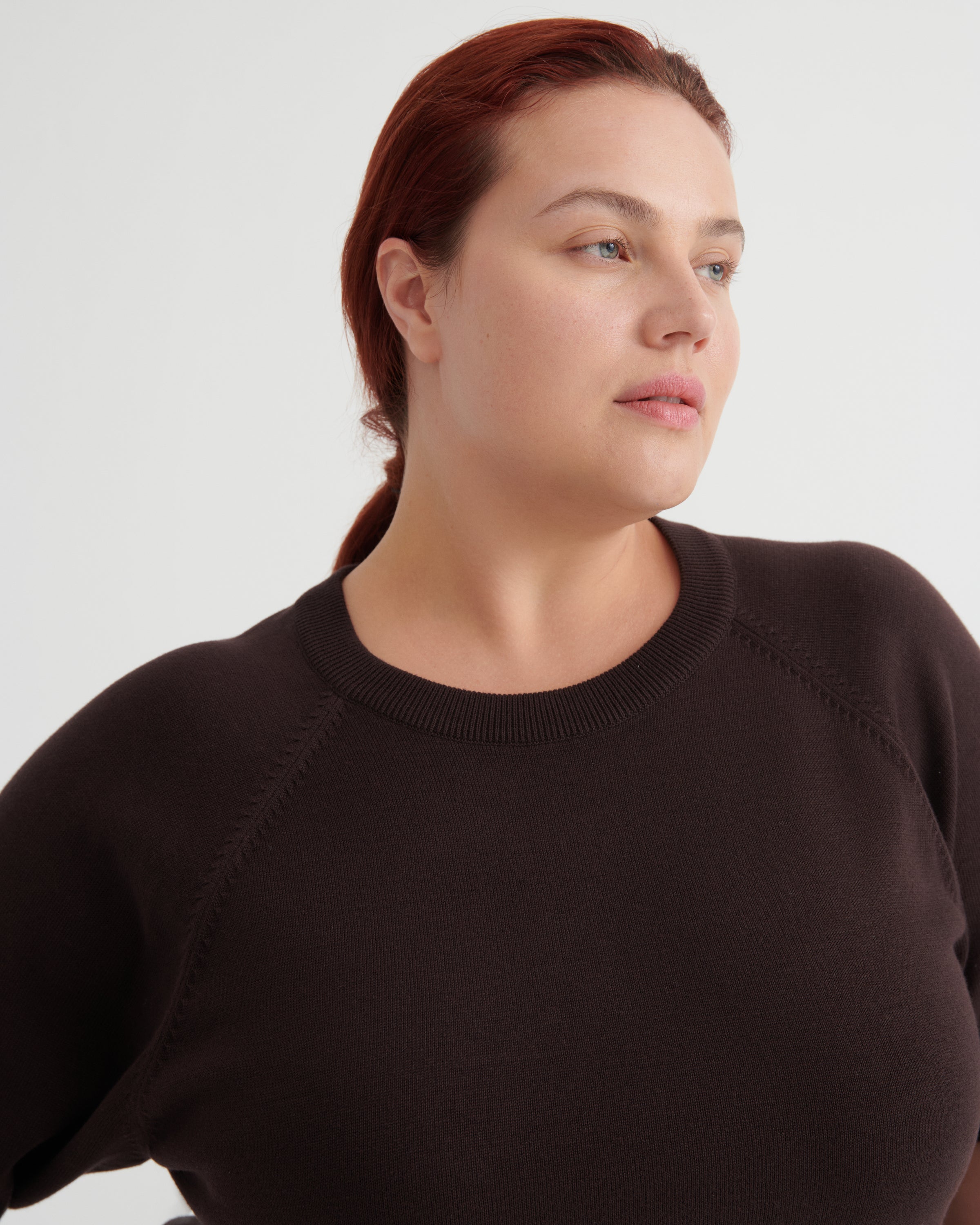 A woman with red hair tied back, wearing a dark brown crewneck knit sweater, shown from the chest up against a light gray studio background.