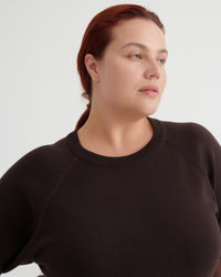 A woman with red hair tied back, wearing a dark brown crewneck knit sweater, shown from the chest up against a light gray studio background.