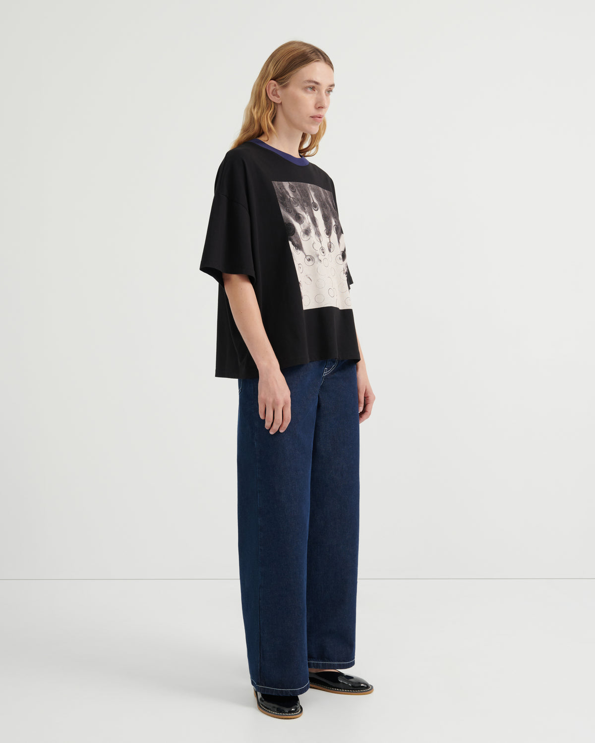 A model stands in profile wearing a black oversized graphic T-shirt with a large black-and-white print on the front, paired with wide-leg dark blue jeans and black shoes, against a clean light gray studio background.