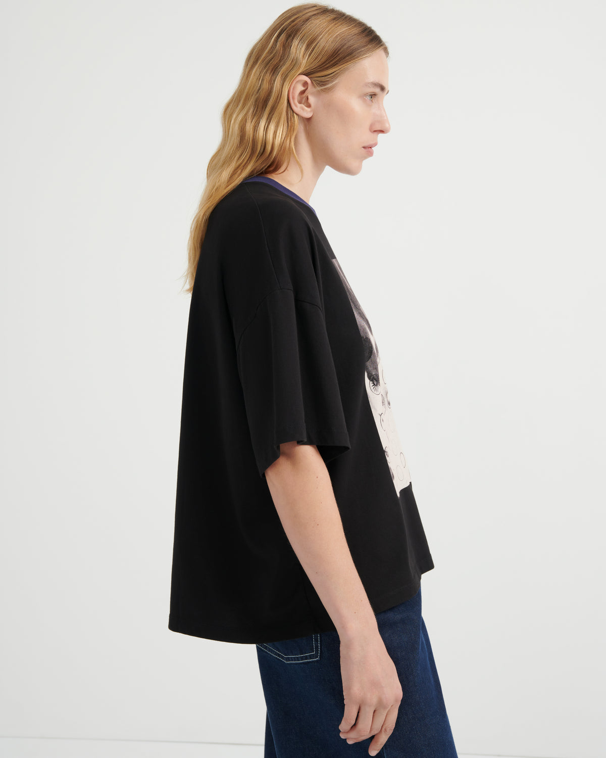 A woman shown in side profile wearing an oversized black short-sleeve T-shirt with a large graphic print on the front, paired with dark blue jeans, against a plain white studio background.