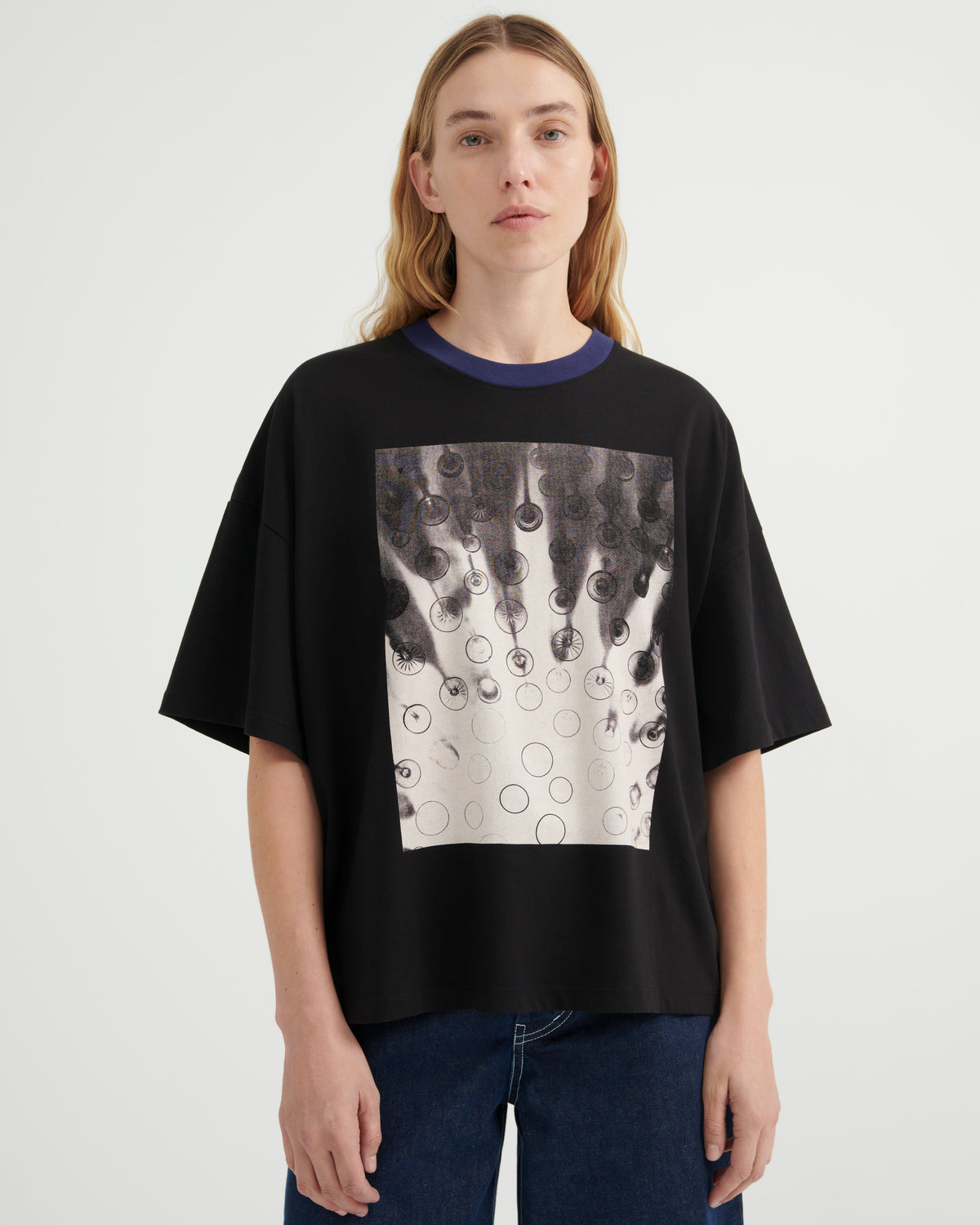 A model wearing a black oversized short-sleeve t-shirt with a dark blue ribbed crew neck and a large square graphic print on the front featuring black and white dripping circles. She is wearing blue jeans against a plain light gray studio background.