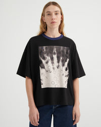 A model wearing a black oversized short-sleeve t-shirt with a dark blue ribbed crew neck and a large square graphic print on the front featuring black and white dripping circles. She is wearing blue jeans against a plain light gray studio background.