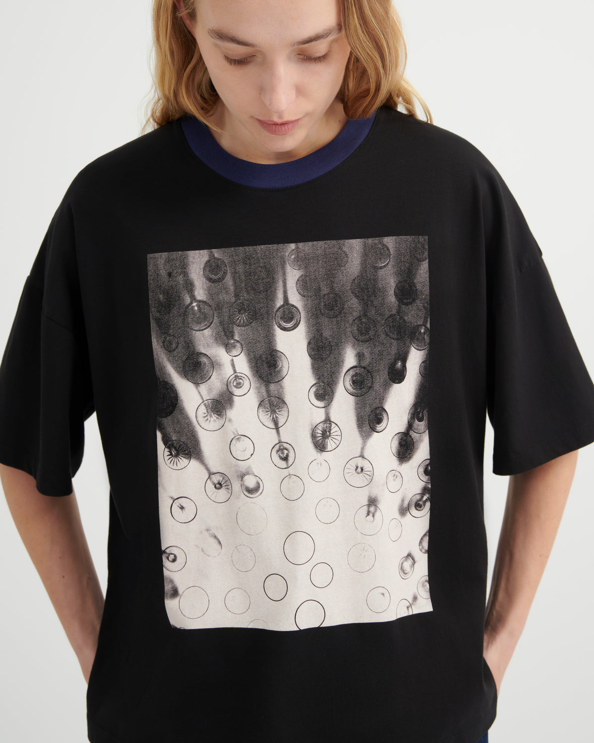A woman wearing an oversized black ringer t-shirt with a dark navy blue crew neck and a large square graphic print on the front featuring black and white bubble patterns.
