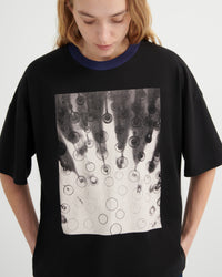 A woman wearing an oversized black ringer t-shirt with a dark navy blue crew neck and a large square graphic print on the front featuring black and white bubble patterns.