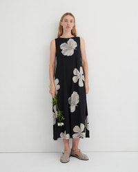 A woman stands against a plain white studio wall wearing a sleeveless black midi dress with a large white floral print. She is holding a small green sprig and wearing flat tan sandals.