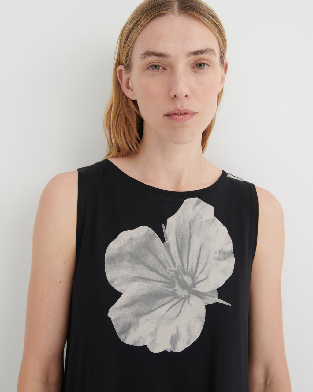 A woman wearing a sleeveless black top with a large gray watercolor-style hibiscus flower graphic on the front, photographed against a plain light gray background.