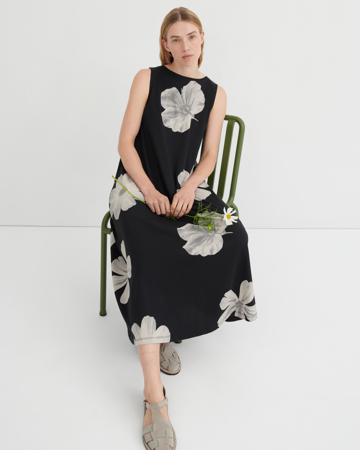 A woman sits on a green metal chair wearing a sleeveless black midi dress with a large white floral print. She is holding a small bouquet of green stems and white daisies against a clean white studio background.