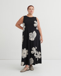 A woman modeling a sleeveless black maxi dress with a large white floral print, paired with gray sandals, against a plain white studio background.