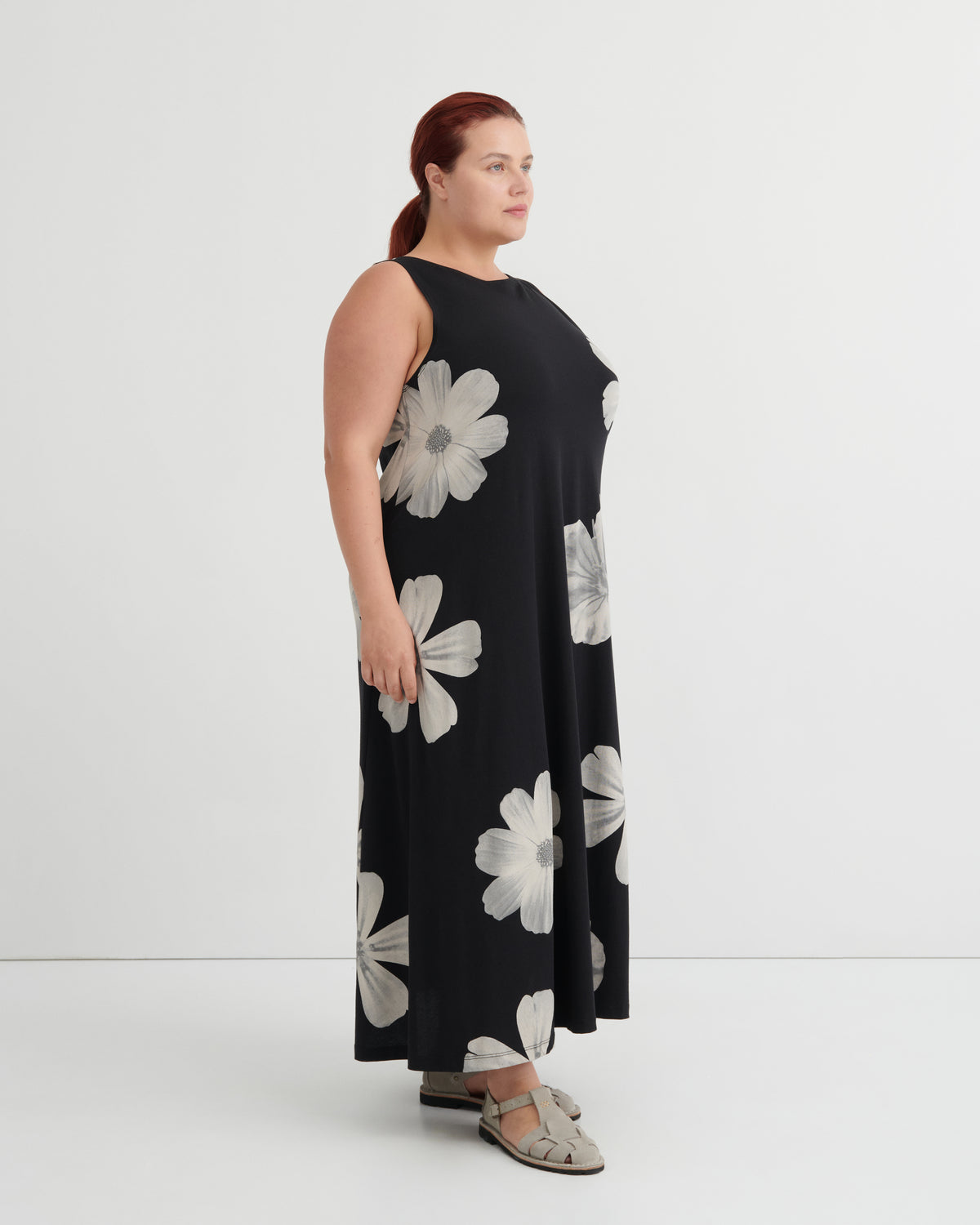 A woman modeling a sleeveless black maxi dress with a large white floral print, shown from a side profile against a plain white studio background.