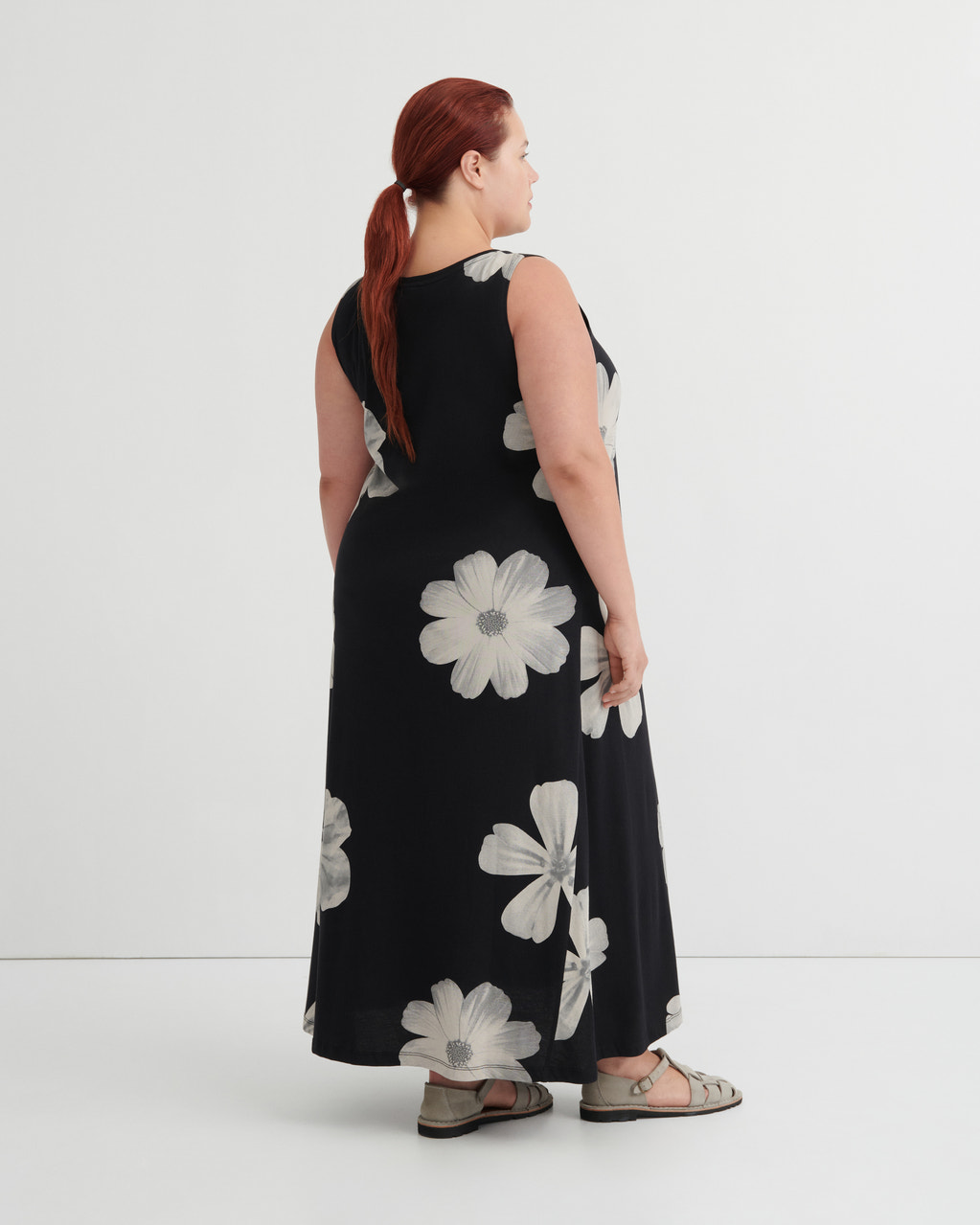 A woman with red hair tied back in a ponytail is shown from the back wearing a sleeveless black maxi dress with a large white floral pattern, paired with gray sandals, against a plain white studio background.
