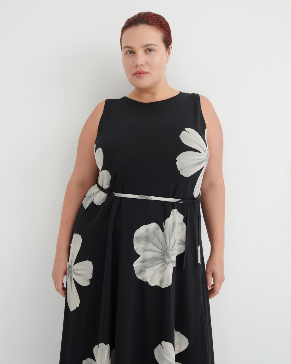 A woman modeling a sleeveless black midi dress with a silver waist tie and large white floral print, set against a plain light gray background.