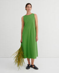 A woman stands against a plain light gray studio background wearing a sleeveless, knee-length bright green dress and dark slip-on shoes, while holding a bunch of yellow-green stems.