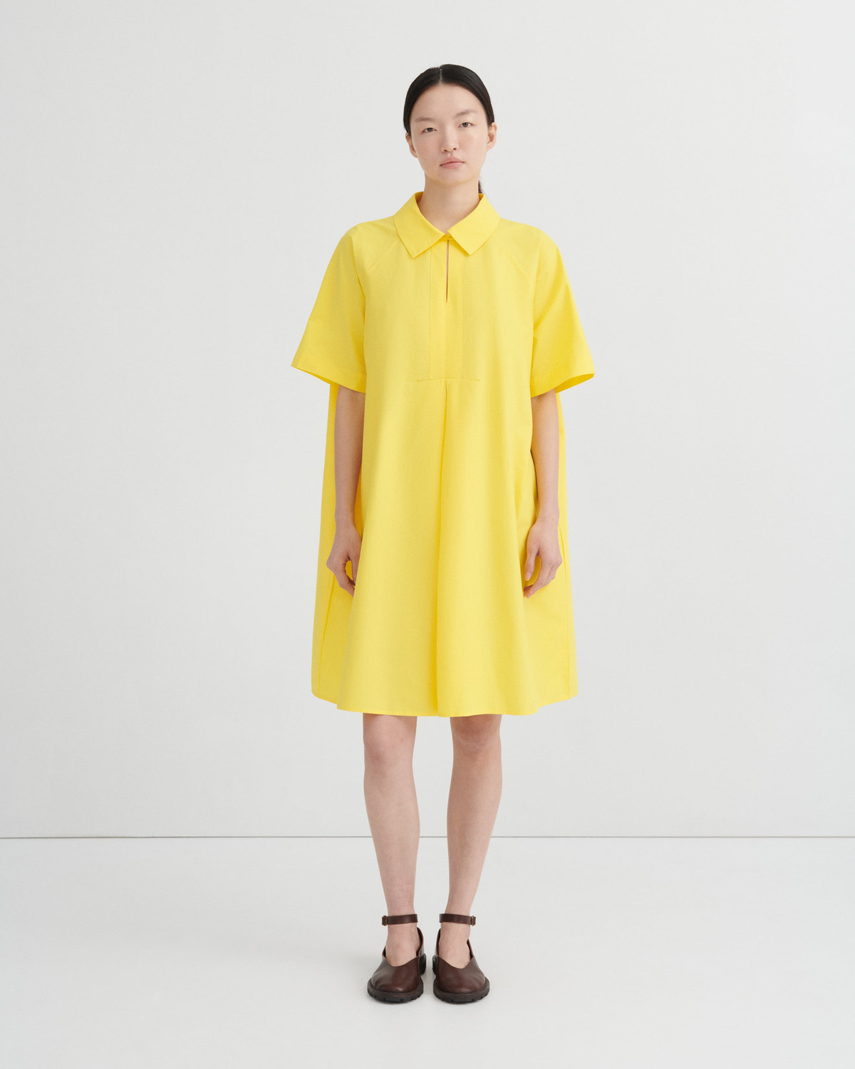 A model stands against a light gray studio background wearing a bright yellow short-sleeve collared dress with a relaxed, oversized fit and dark brown Mary Jane shoes.