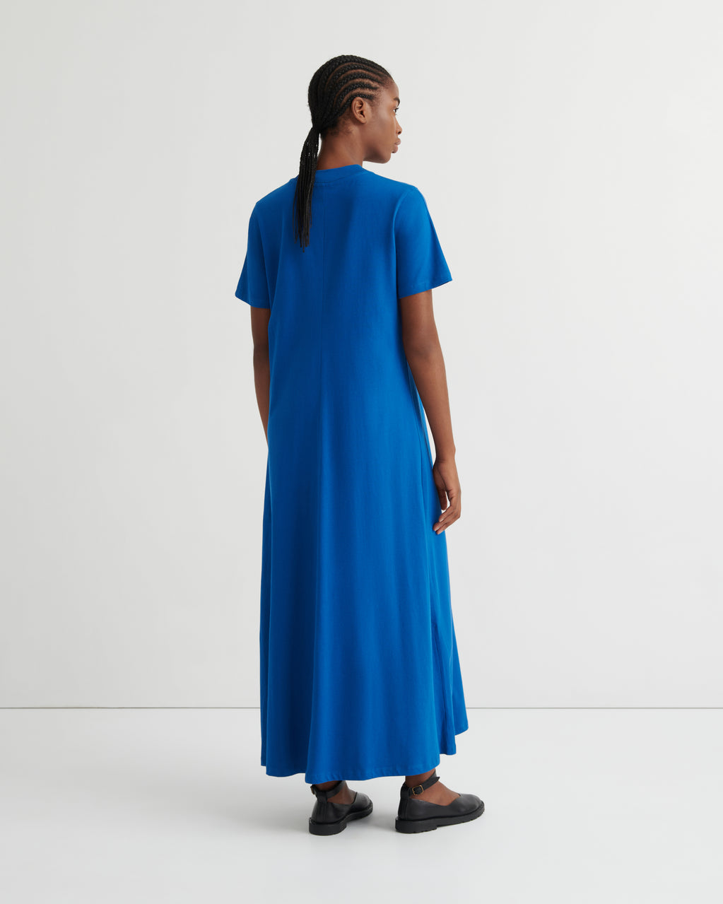 A woman wearing a bright blue short-sleeve maxi dress, shown from the back in a studio setting with a white background.