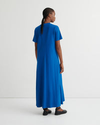 A woman wearing a bright blue short-sleeve maxi dress, shown from the back in a studio setting with a white background.