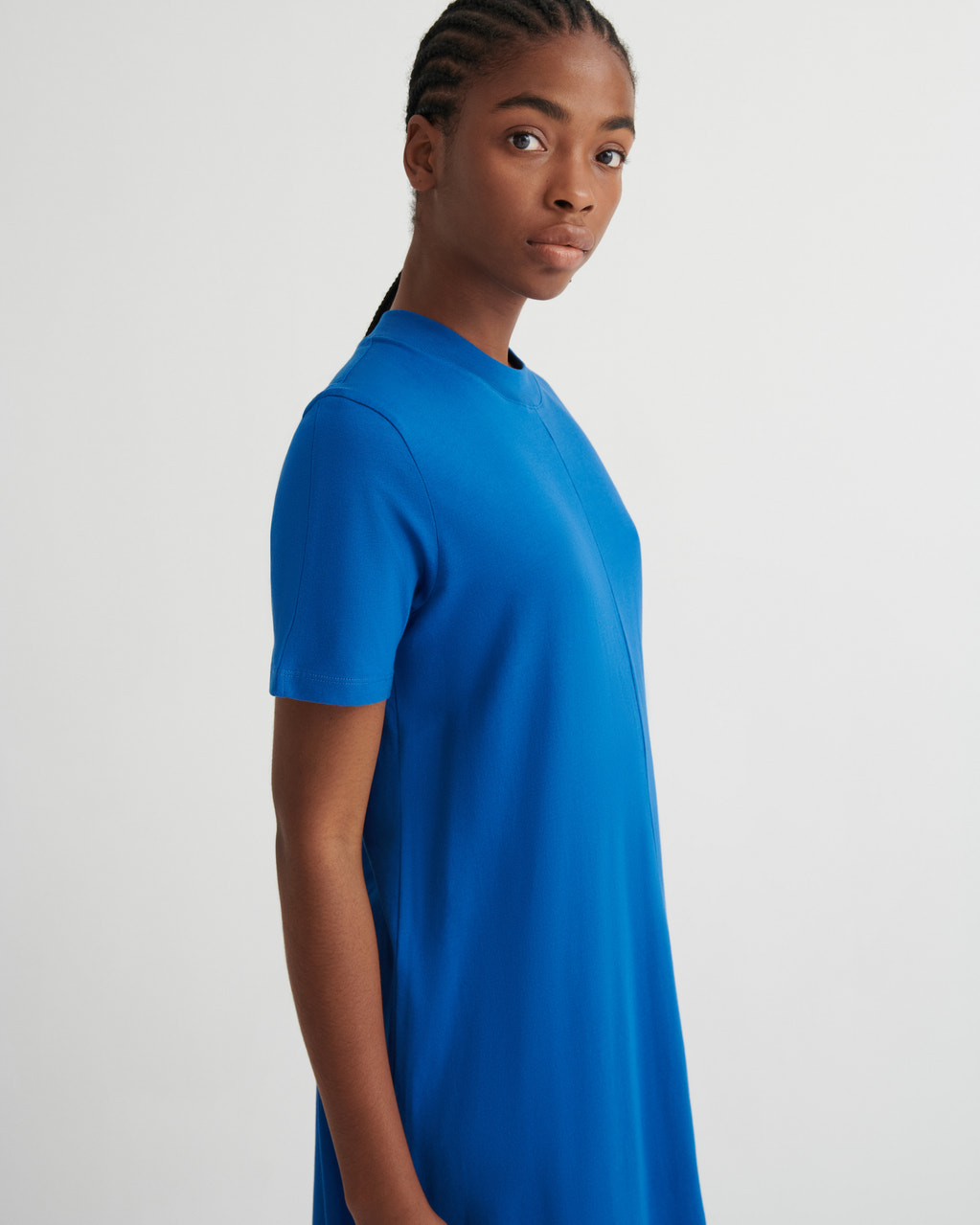 A woman wearing a bright blue short-sleeve t-shirt dress, shown from a side profile against a light gray studio background.