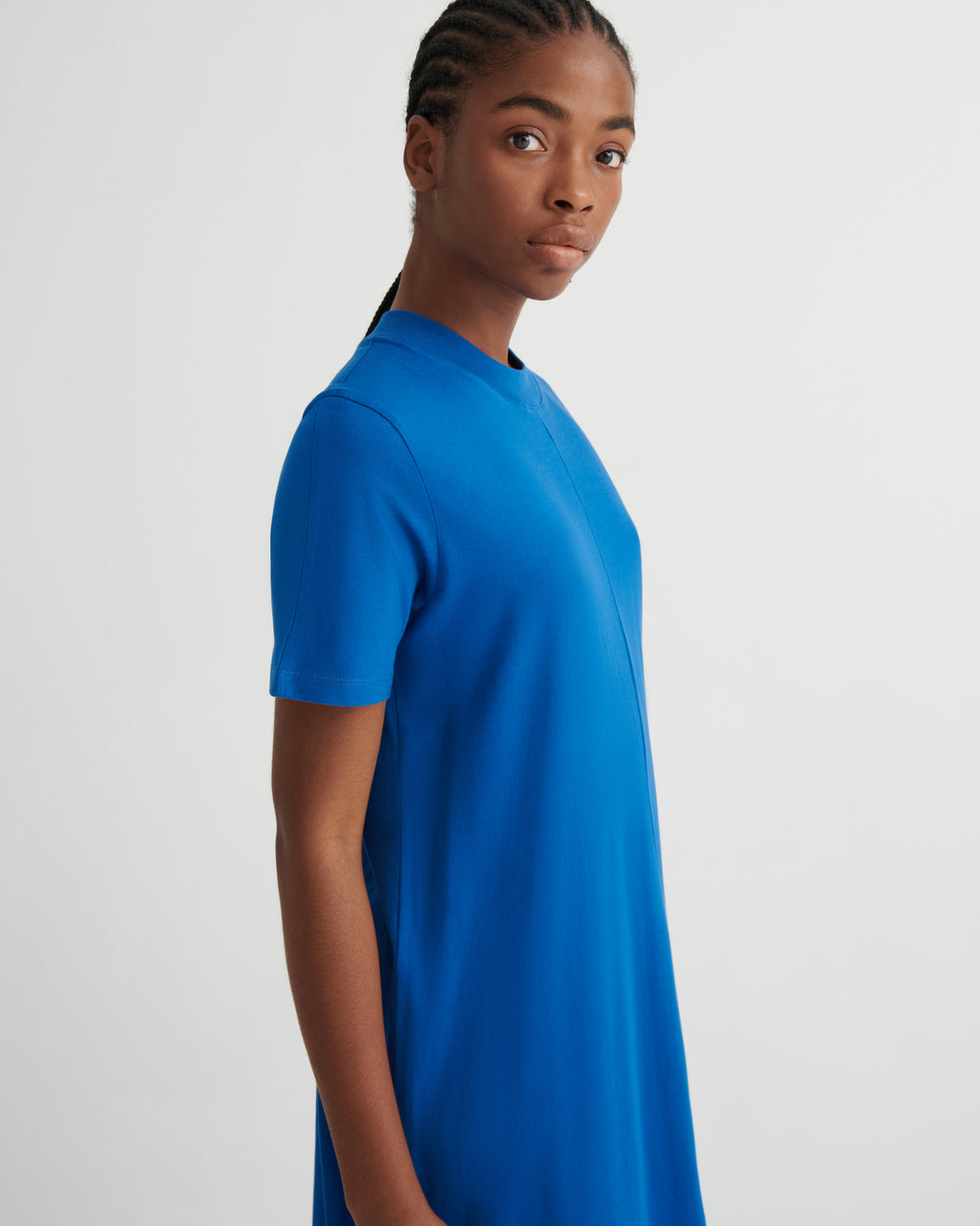A woman wearing a bright blue short-sleeve t-shirt dress, shown from a side profile against a light gray studio background.
