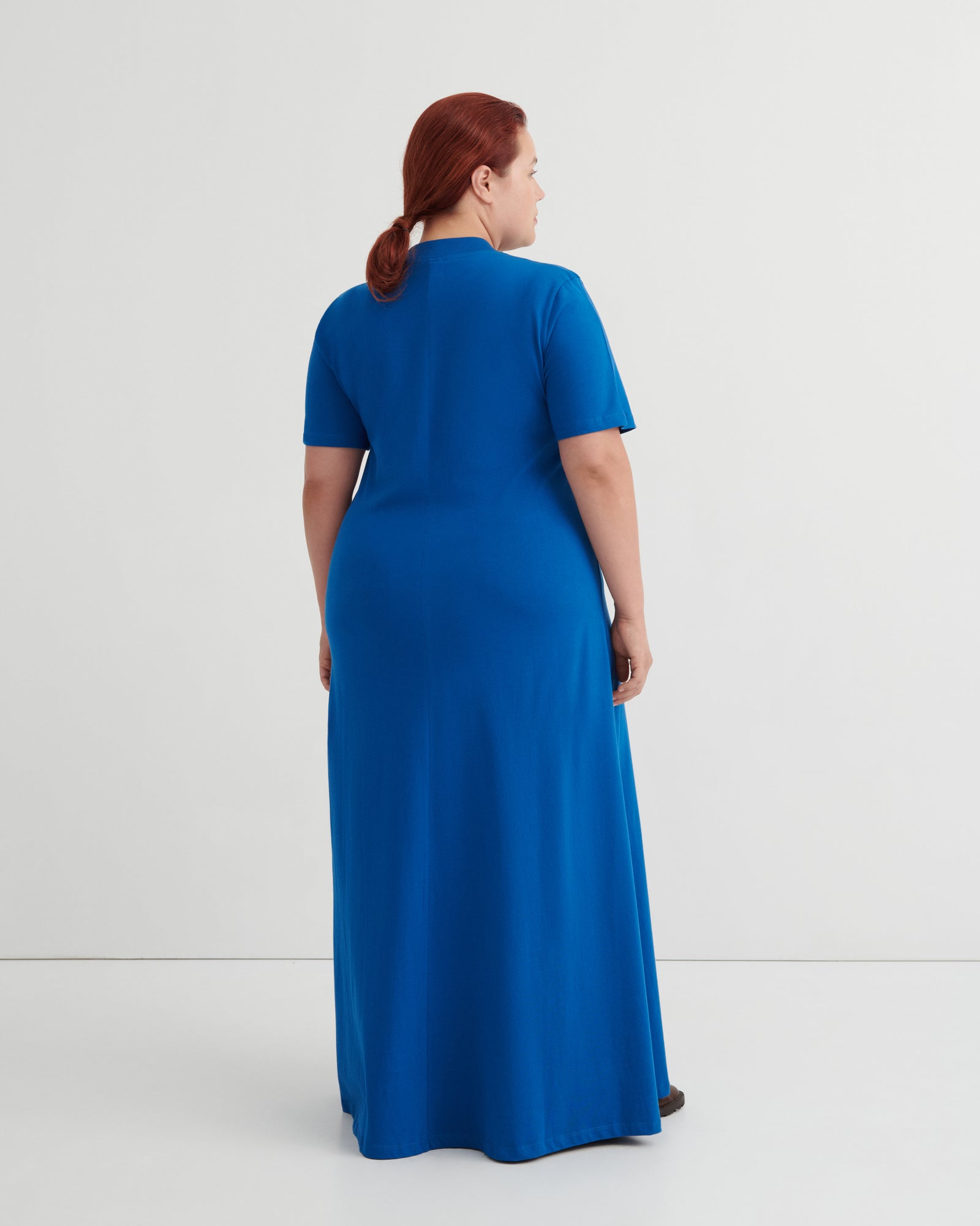 A woman with red hair tied back is shown from the back wearing a bright blue short-sleeve maxi dress against a plain light gray studio background.