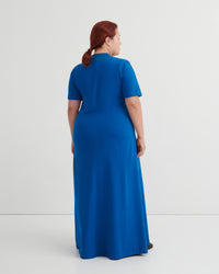 A woman with red hair tied back is shown from the back wearing a bright blue short-sleeve maxi dress against a plain light gray studio background.