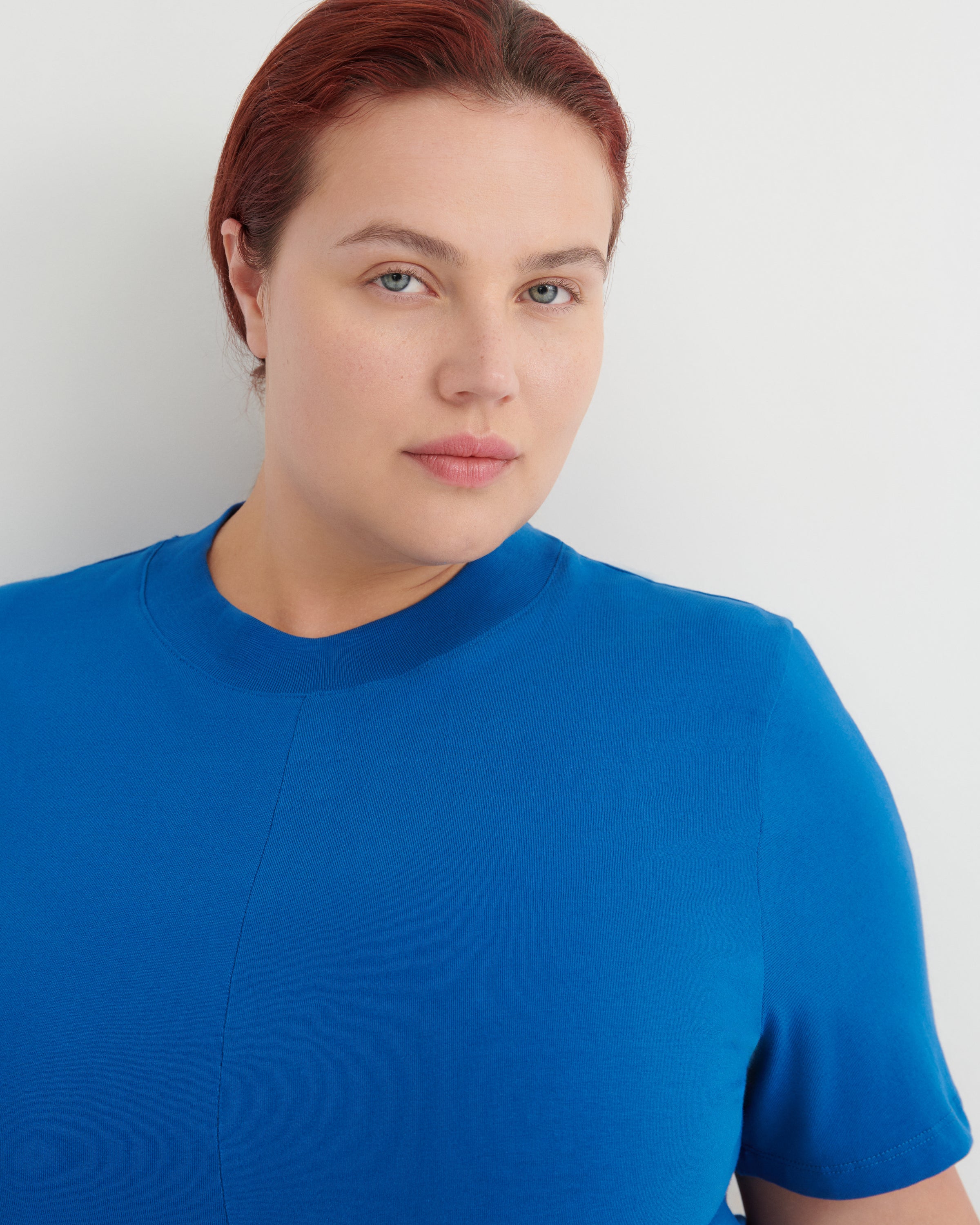 A close-up shot of a woman with red hair wearing a bright blue short-sleeve crewneck top against a light gray background.