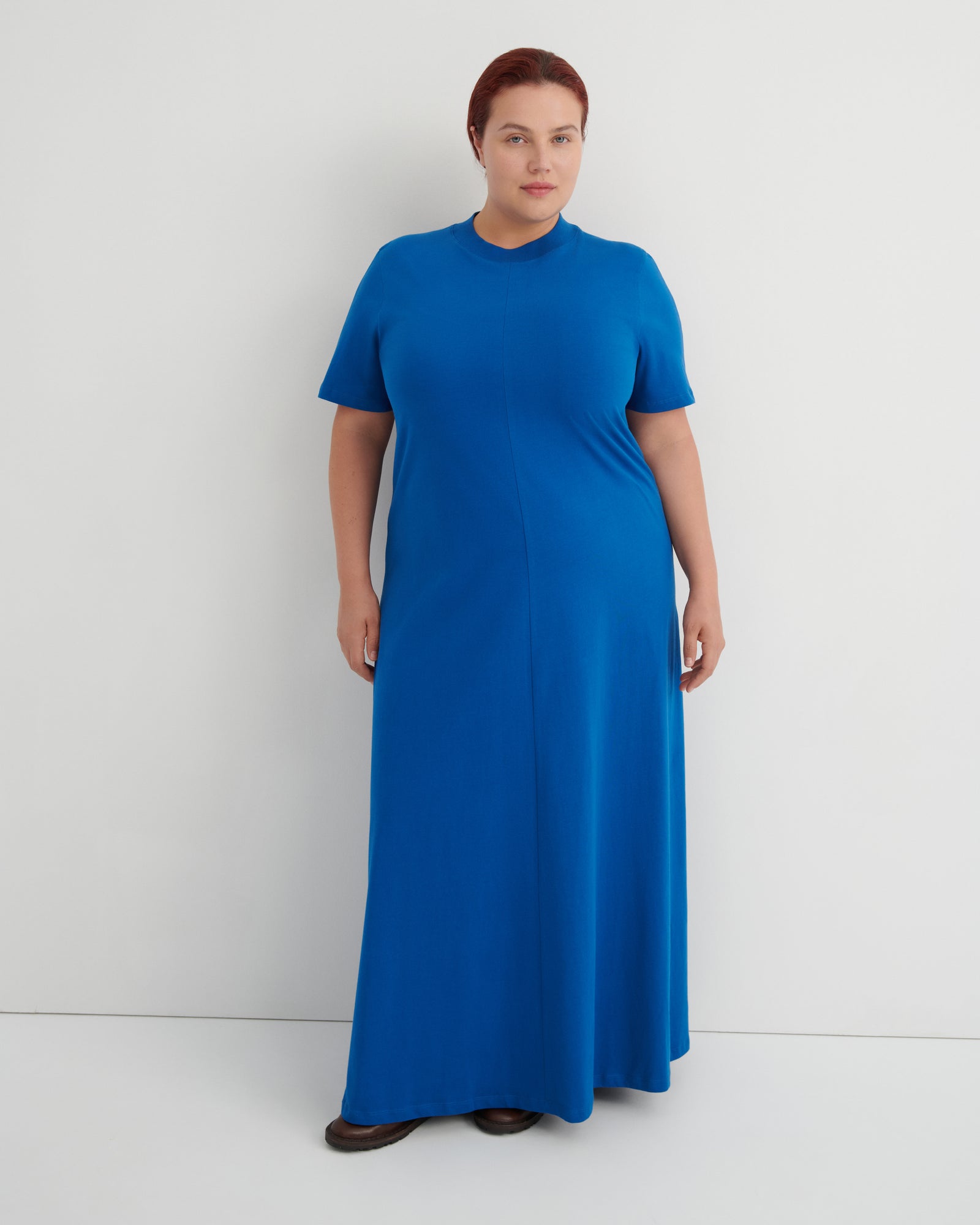 A woman modeling a bright blue short-sleeve maxi dress with a mock neck, standing against a plain light gray studio background.