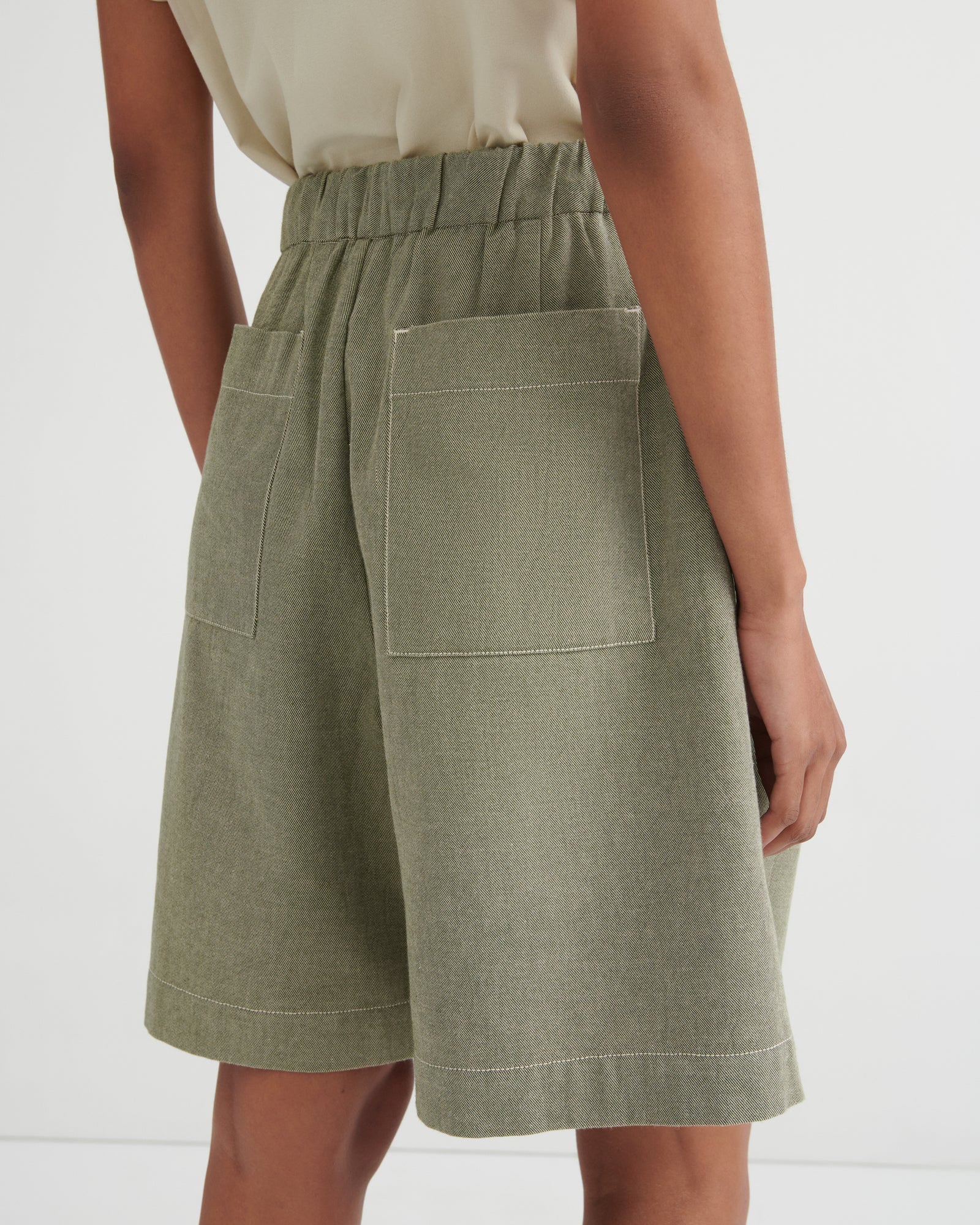 A close-up back view of a model wearing sage green linen shorts with an elastic waistband and large patch pockets, paired with a light beige top against a white studio background.
