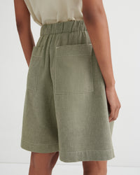 A close-up back view of a model wearing sage green linen shorts with an elastic waistband and large patch pockets, paired with a light beige top against a white studio background.