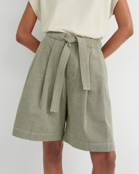 A person wearing high-waisted olive green pleated shorts with a matching tie belt, paired with a cream sleeveless top, against a light gray studio background.