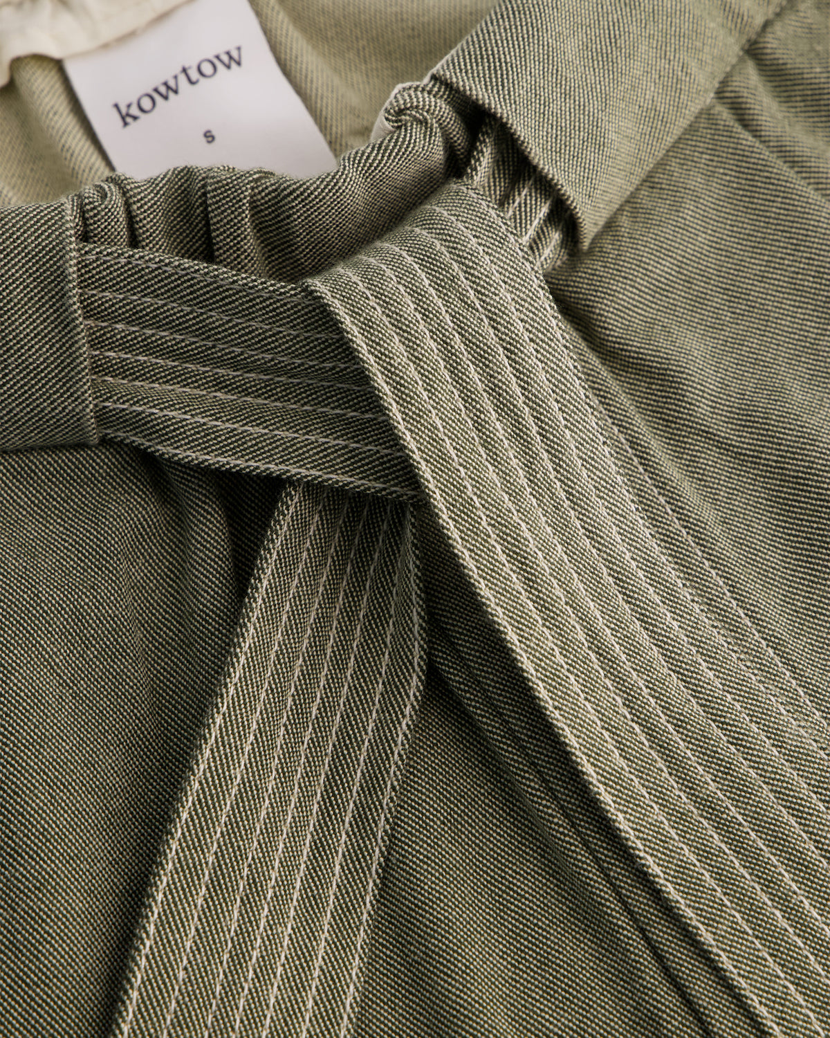Close-up of a Kowtow garment in olive green fabric with a matching tie belt and a white brand label visible at the waistband.